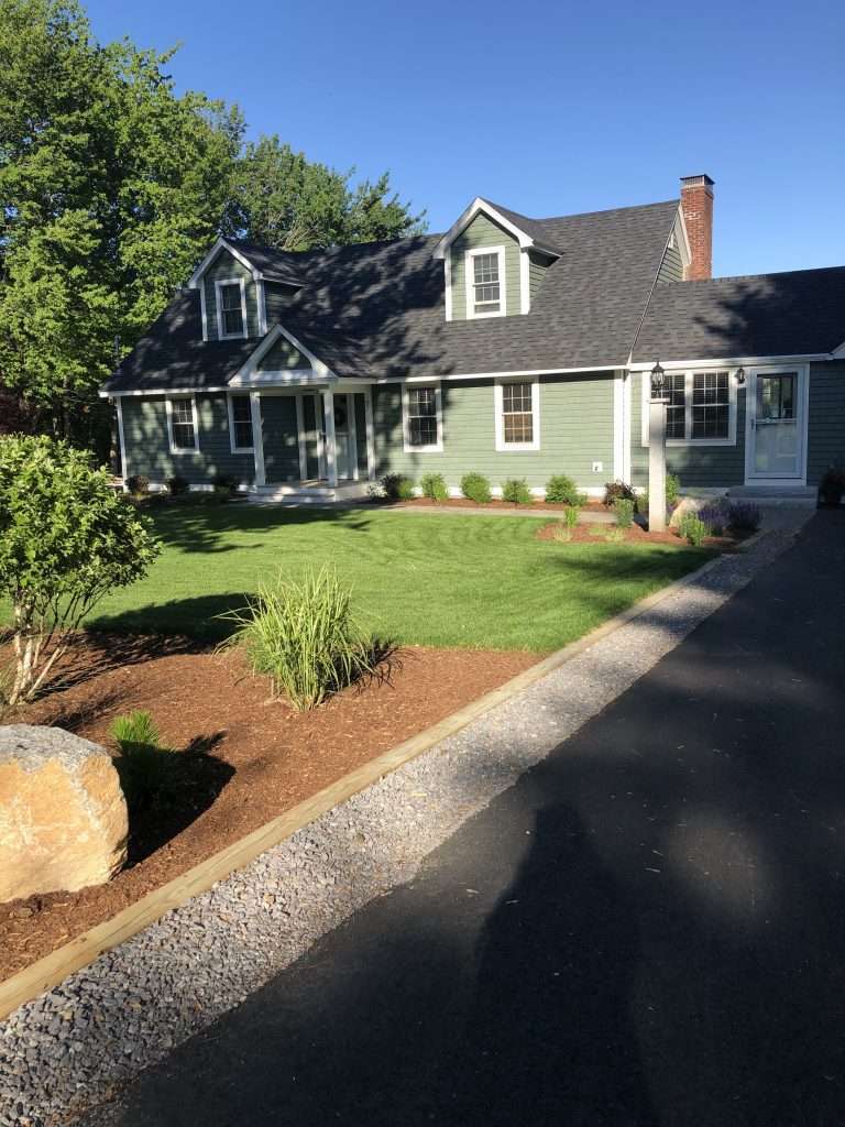Professional foundation planting and front yard landscape installation featuring fresh mulch beds, ornamental grasses, flowering shrubs, granite accents, and manicured lawn surrounding a New Hampshire home