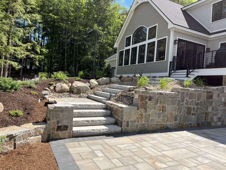 Luxury hardscape installation with custom paver patio, granite stairway, natural stone retaining wall, decorative landscape boulders, and modern outdoor living design for a New Hampshire home