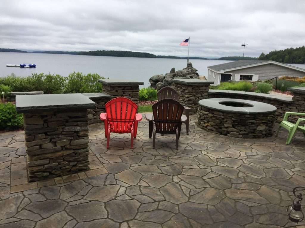 Fire Pit Ideas 2 Natural stone lakeside patio with custom masonry fire pit, stacked stone seating walls, granite caps, Adirondack chairs, and panoramic waterfront views designed for outdoor entertaining and relaxation