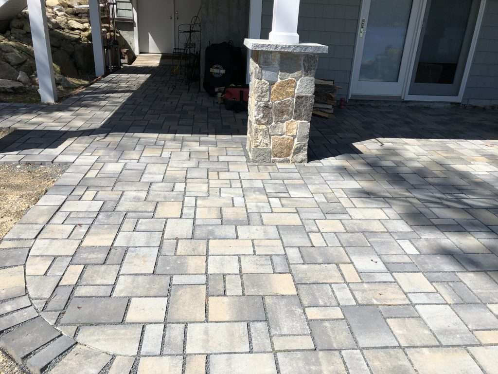 Custom paver patio and covered entryway installation featuring multi-pattern stone pavers, curved border detail, and natural stone column veneer in New Hampshire