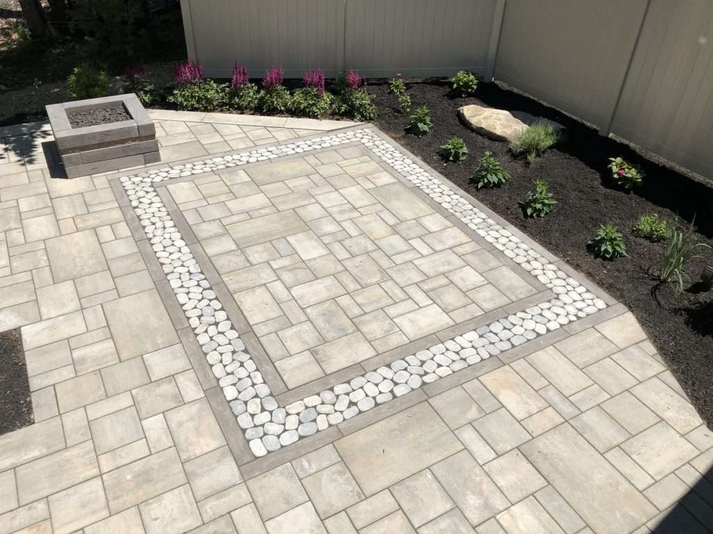 Custom paver patio with decorative river rock inlay, modern fire pit, geometric hardscape design, and landscaped garden beds for a stylish backyard outdoor living space
