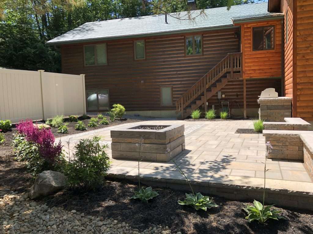 Backyard patio with custom paver hardscape, modern fire pit, built-in outdoor kitchen, retaining wall seating, granite steps, and professionally landscaped garden beds beside a rustic log home
