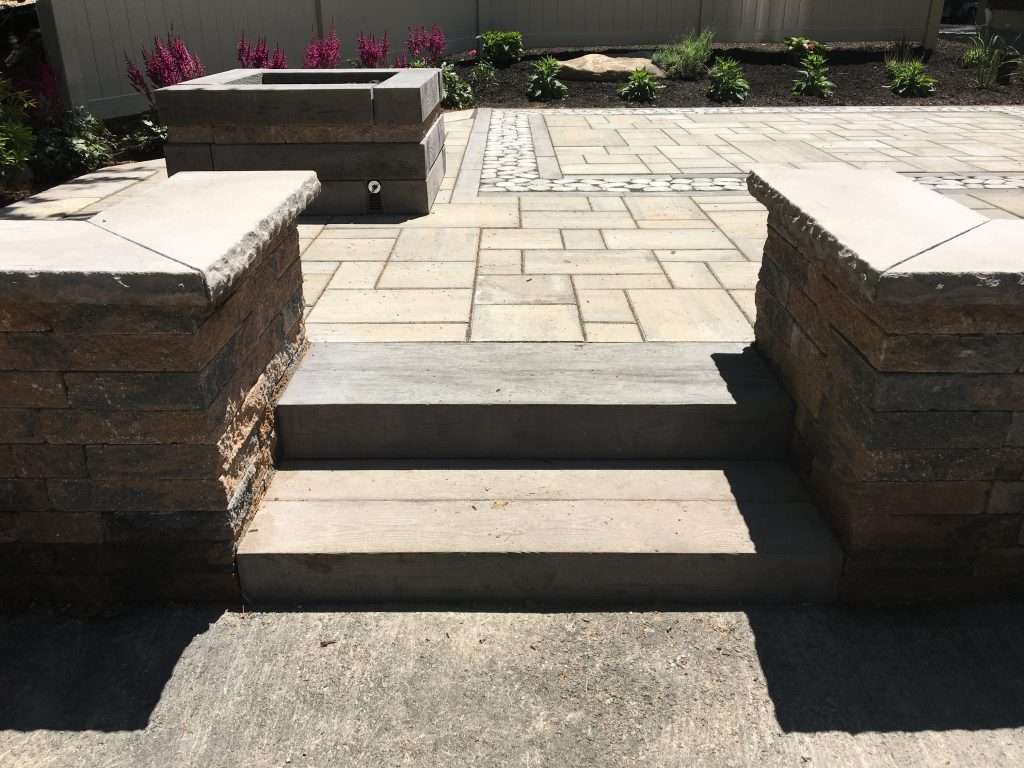 Custom paver patio installation featuring architectural stone steps, seating walls, modern fire pit, decorative stone inlays, and professionally designed outdoor living space in New Hampshire