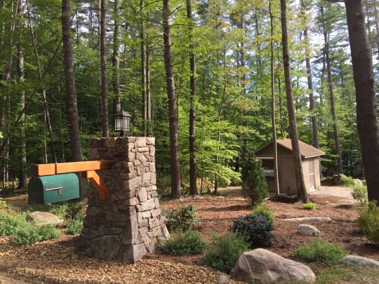 Custom natural stone mailbox column with integrated lighting, decorative river rock, woodland landscaping, native plantings, and rustic curb appeal enhancements for a New Hampshire property