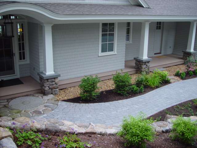Curved paver walkway with natural stone accents, decorative foundation plantings, river rock beds, and custom front entry landscaping designed to enhance curb appeal for a New Hampshire home