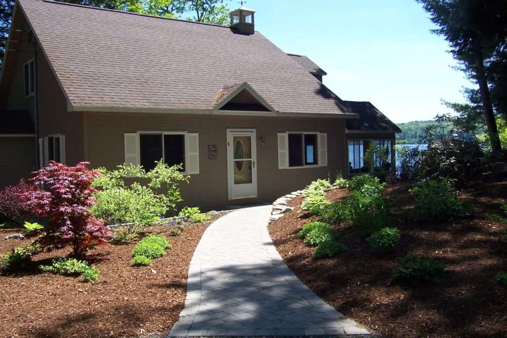 Curved paver walkway and custom landscape design with ornamental trees, foundation plantings, and fresh mulch surrounding a New Hampshire lakeside home